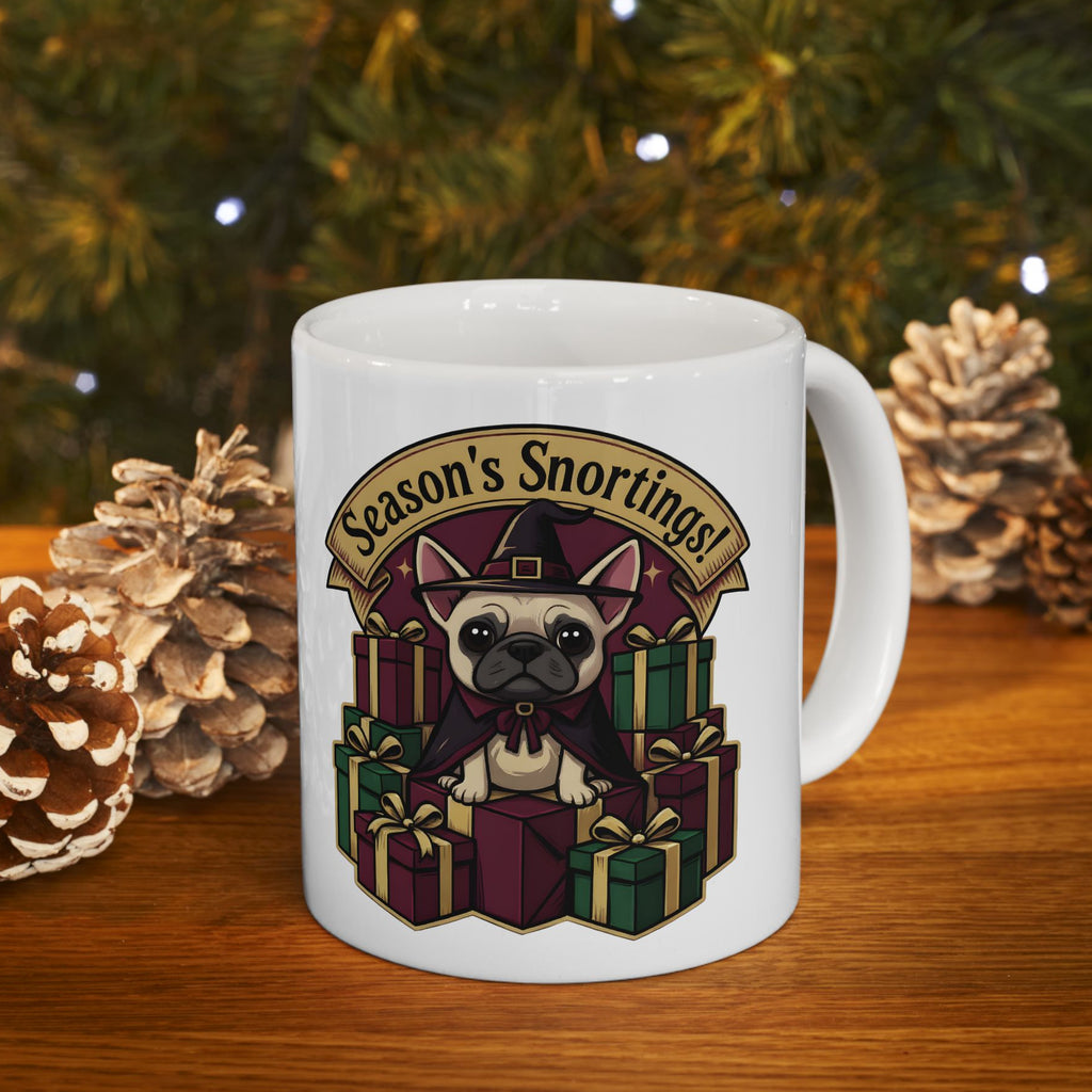 White mug with a dog design and 'Season's Snortings!' text, placed on a wooden surface with pinecones and a Christmas tree in the background.