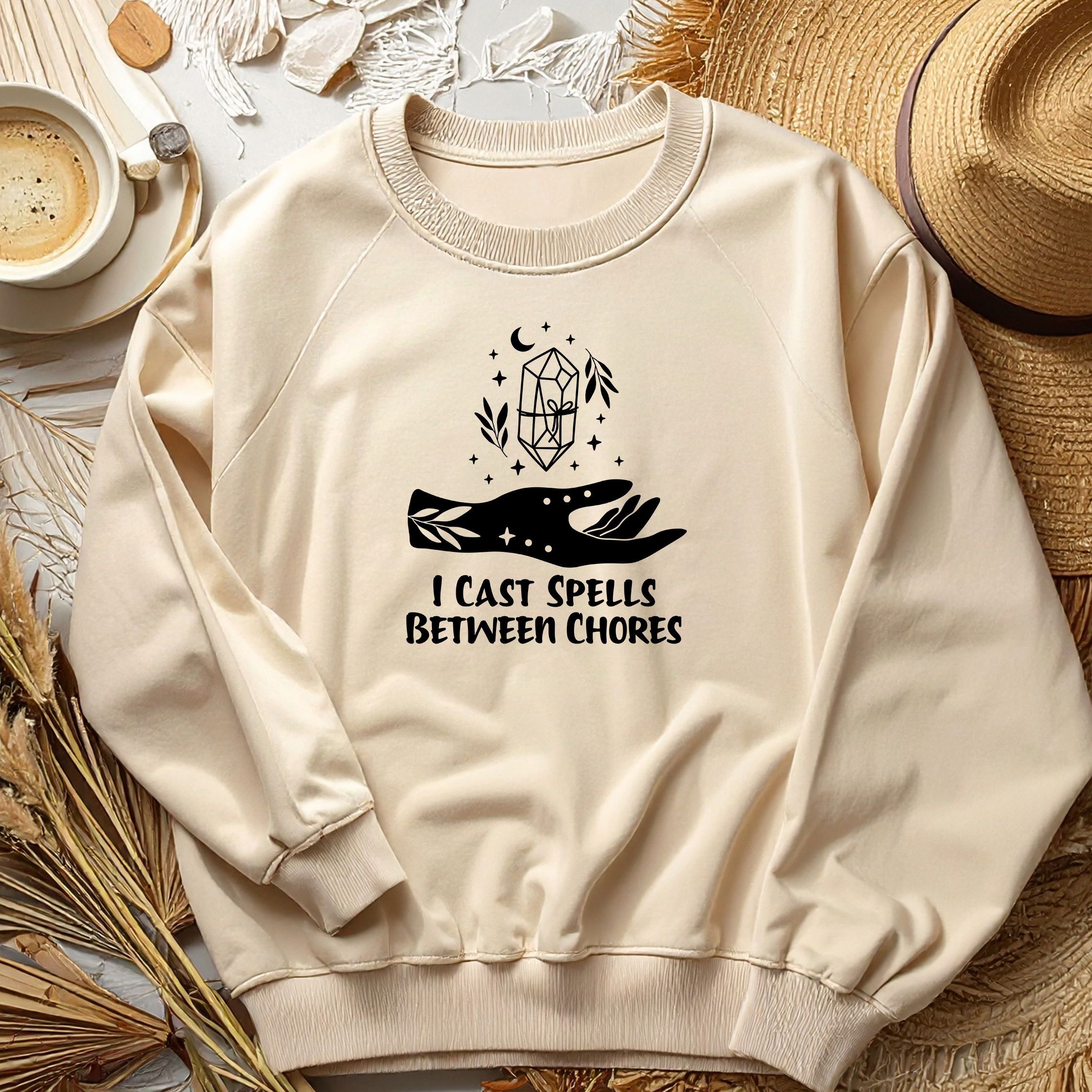 Beige sweatshirt with a graphic design and text, placed on a surface with a cup of coffee and straw hat.