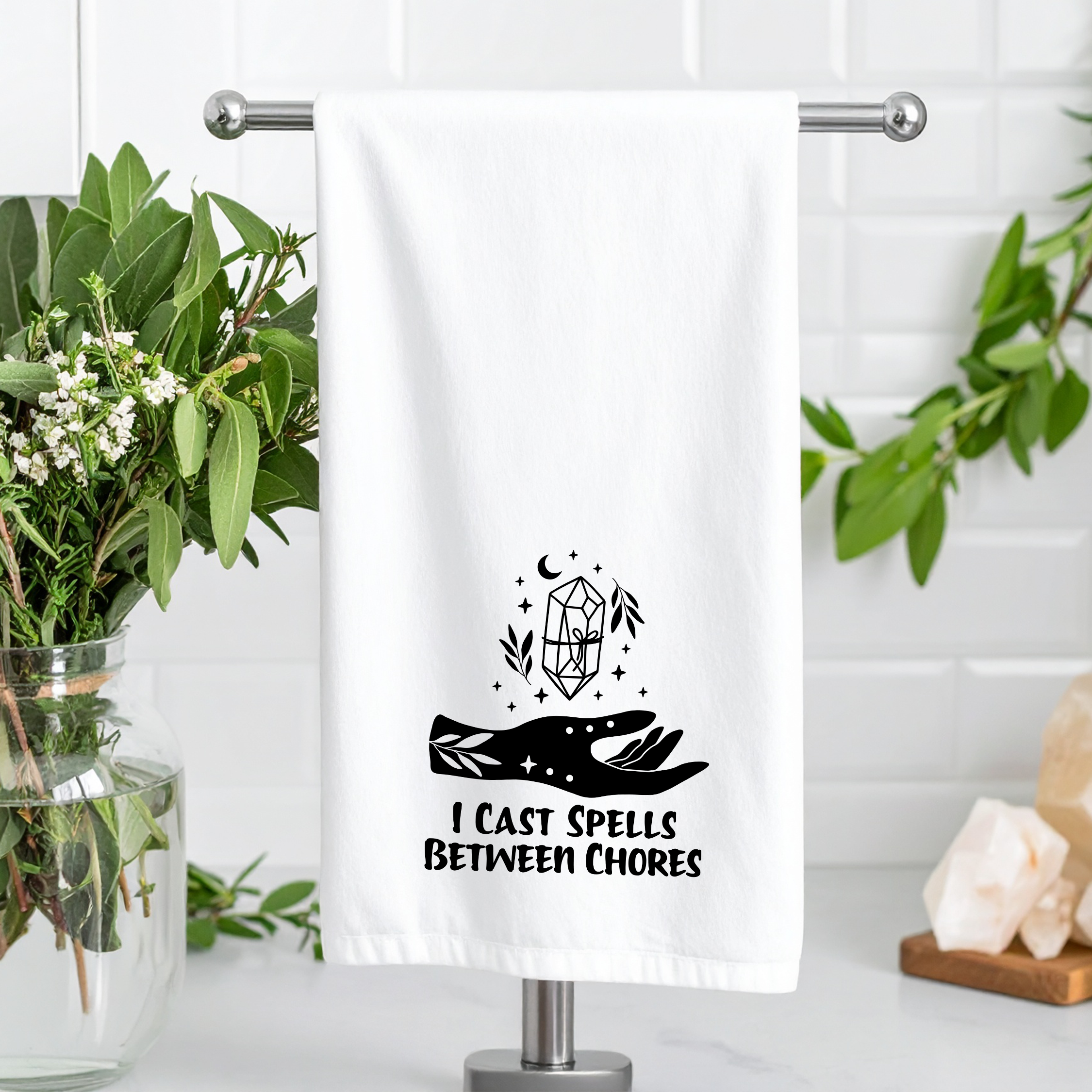 Towel with 'I Cast Spells Between Chores' design on a stand with plants and decor.