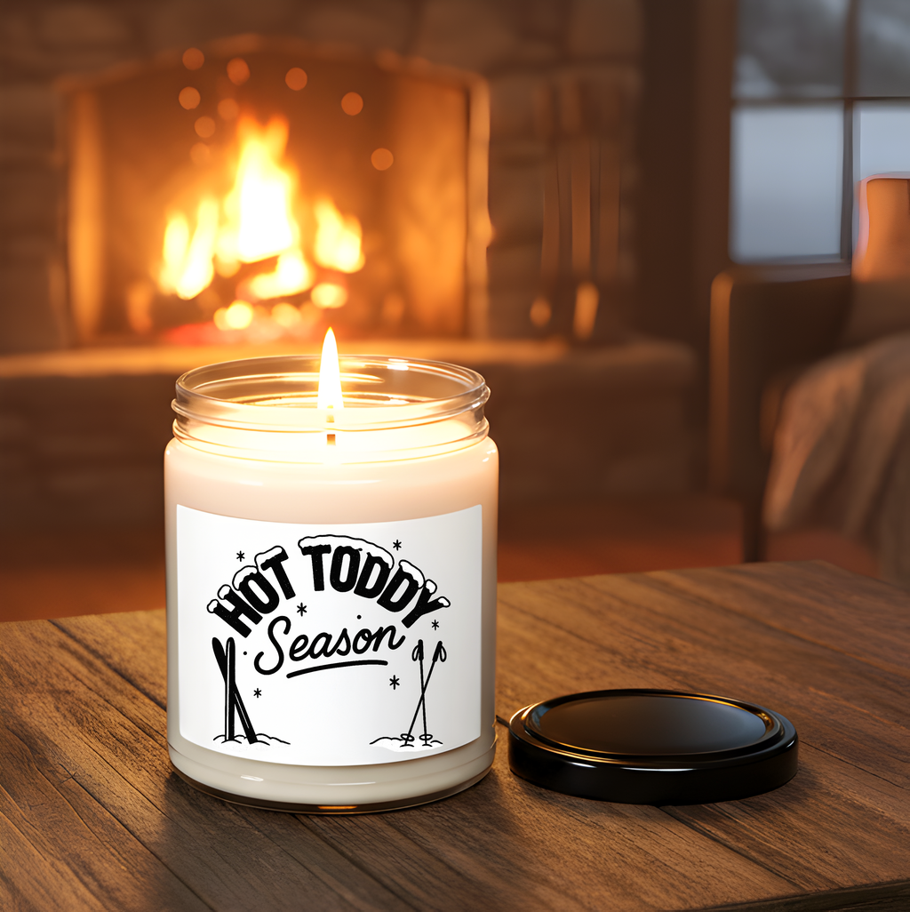 Candle labeled 'Hot Toddy Season' on a wooden surface with a fireplace in the background