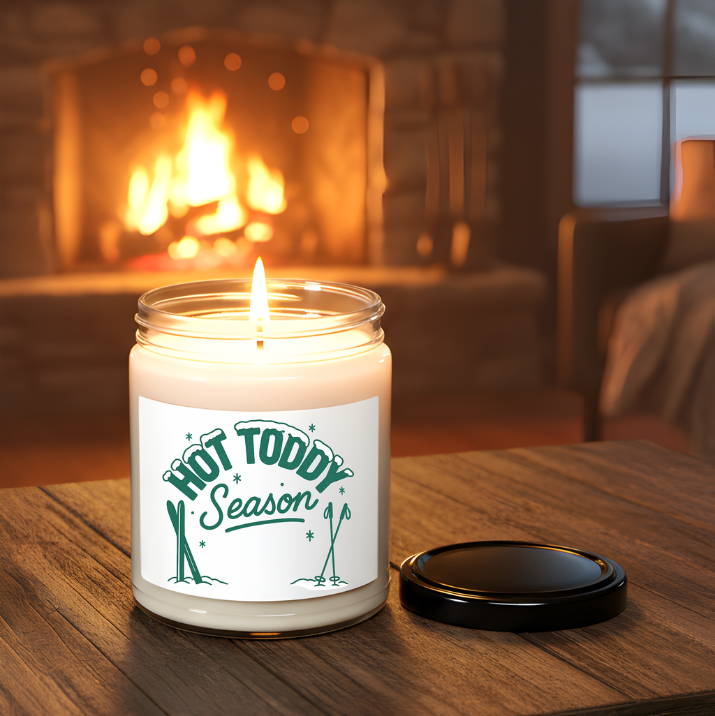 Candle in a jar labeled 'Hot Toddy Season' on a wooden surface with a fireplace in the background